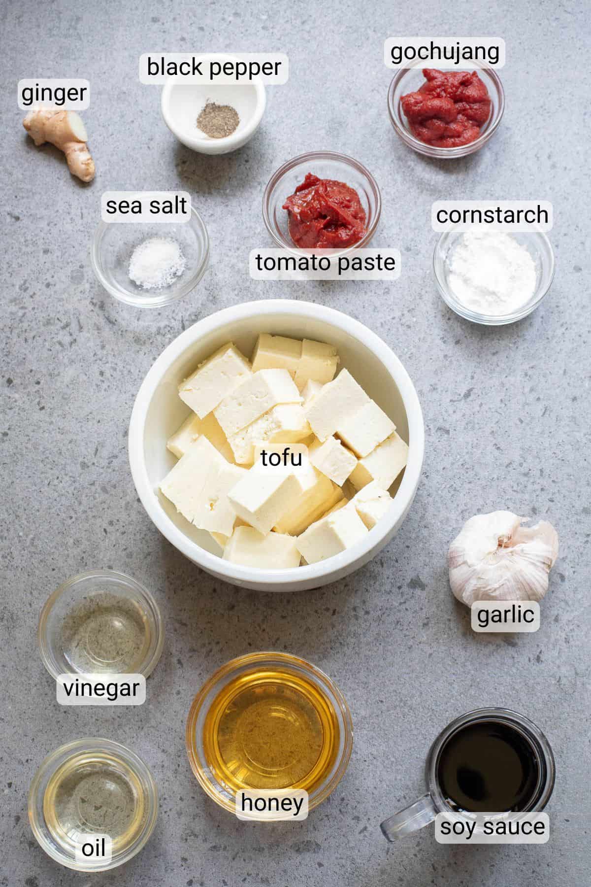 Ingredients for a dish are arranged on a countertop: tofu, ginger, black pepper, gochujang, sea salt, tomato paste, cornstarch, garlic, oil, vinegar, honey, and soy sauce.