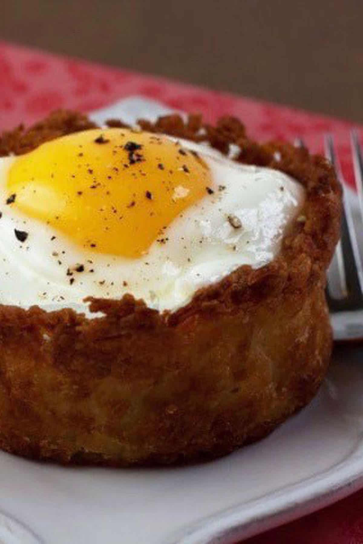 A baked dish with a crispy crust, topped with a sunny-side-up egg and sprinkled with black pepper, on a white plate. A fork is visible in the background.