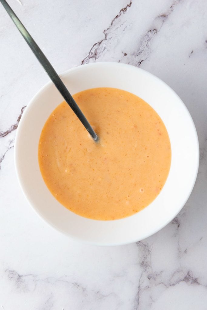 A white bowl filled with creamy orange soup, with a metal spoon resting inside, placed on a white marble surface.