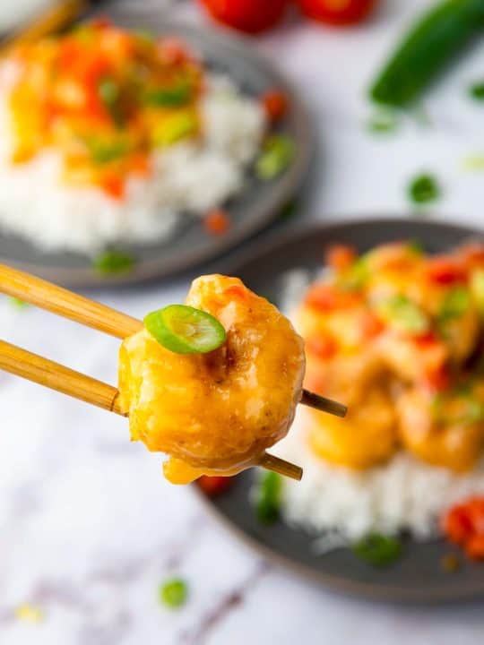 A piece of breaded shrimp held by chopsticks, with two plates of shrimp and rice in the background, garnished with green onions and diced tomatoes.