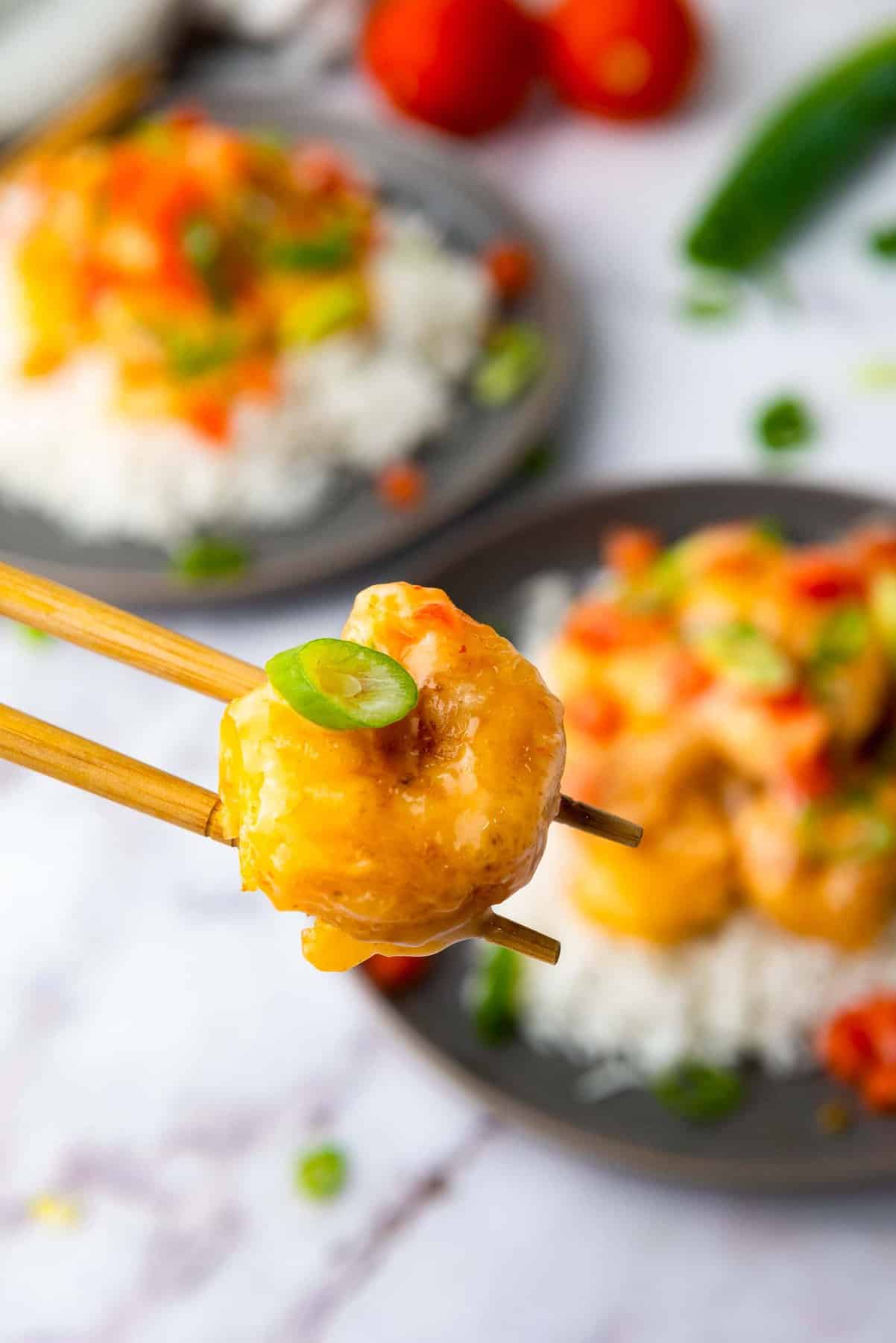 A piece of breaded shrimp held by chopsticks, with two plates of shrimp and rice in the background, garnished with green onions and diced tomatoes.