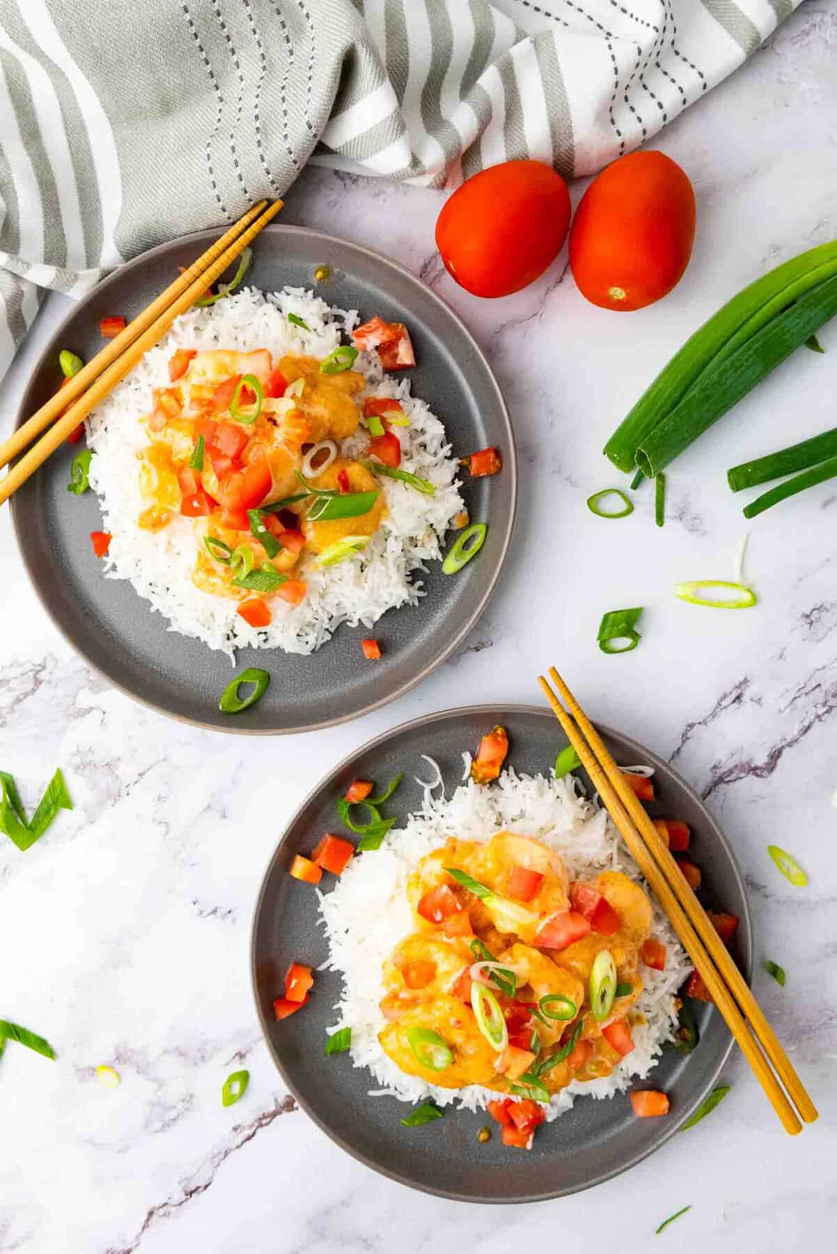 Two plates of rice topped with diced tomatoes, green onions, and a creamy sauce. Chopsticks placed on the plates. Whole tomatoes, a green onion, and a striped cloth nearby on a marble surface.
