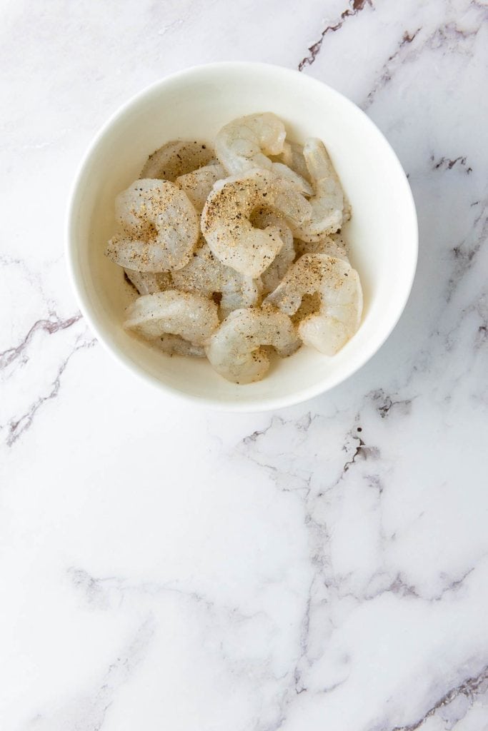 Bowl of raw shrimp seasoned with pepper on a marble surface.