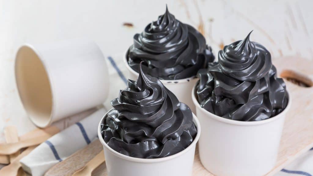 Three cups of soft serve ice cream with black swirls, arranged on a wooden surface with an overturned cup and napkins in the background.