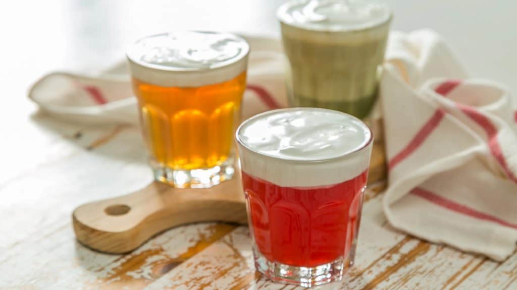 Three glasses of frothy beverages sit on a wooden board. The drinks are amber, red, and green in color, with a striped cloth in the background.