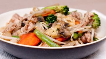A bowl of stir-fried vegetables, including bean sprouts, carrots, broccoli, and mushrooms, mixed with pieces of chicken.