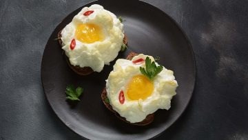 Two cloud eggs with runny yolks on toast, garnished with red chili slices and parsley, served on a black plate.