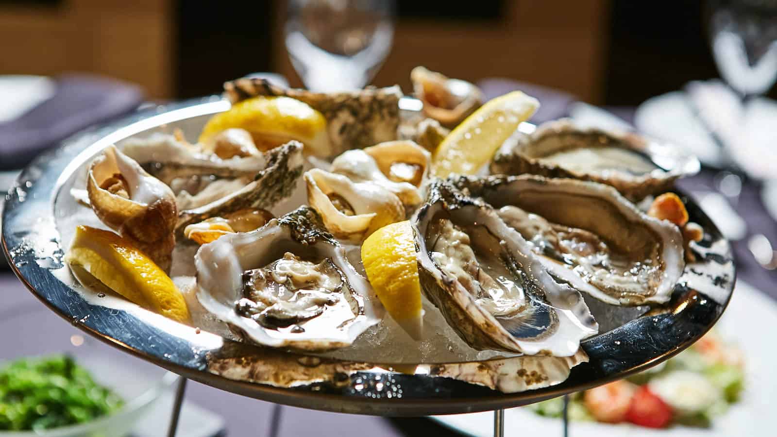 A platter of shucked oysters on ice, garnished with lemon wedges, served on a metal stand.