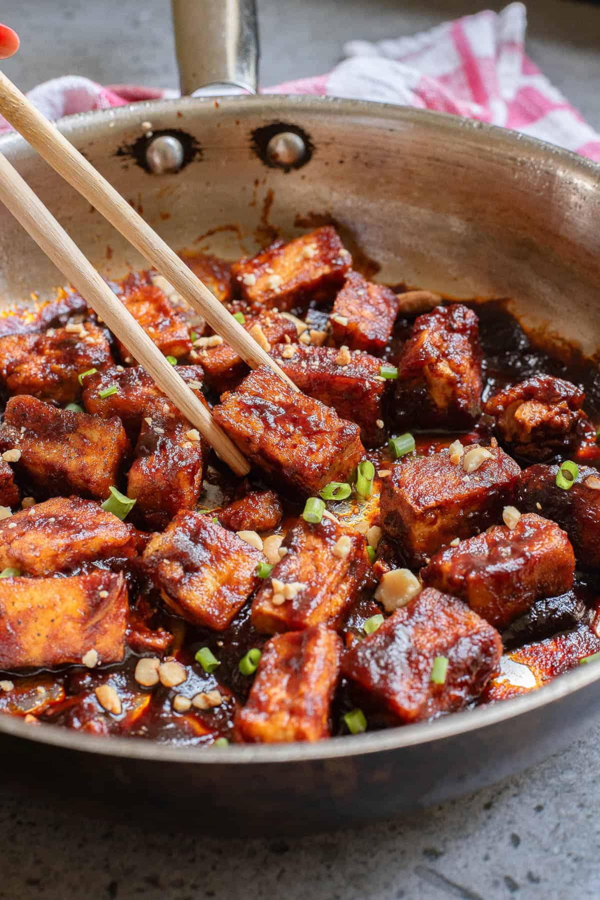 A skillet filled with cooked tofu cubes in a dark sauce, topped with chopped green onions and sesame seeds. A pair of chopsticks is poised to pick up a piece.