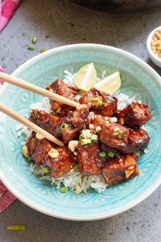 A bowl of rice topped with glazed tofu pieces, garnished with chopped peanuts and green onions. Two lime wedges and chopsticks are placed on top.