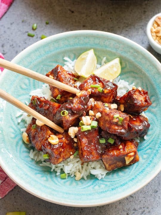 A bowl of rice topped with glazed tofu pieces, garnished with chopped peanuts and green onions. Two lime wedges and chopsticks are placed on top.