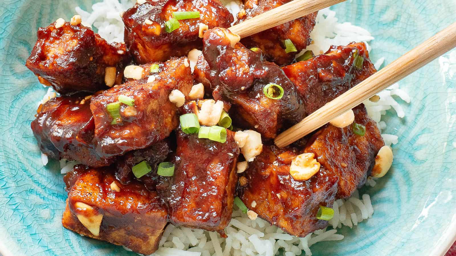 A bowl of glazed tofu pieces garnished with chopped scallions and peanuts, served over white rice with chopsticks.