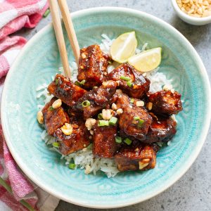 A bowl of rice topped with glazed tofu cubes, garnished with peanuts and green onions, served with lime wedges.