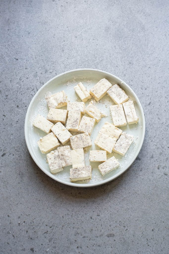 A white plate containing several pieces of cubed tofu, seasoned with pepper, resting on a gray speckled countertop.