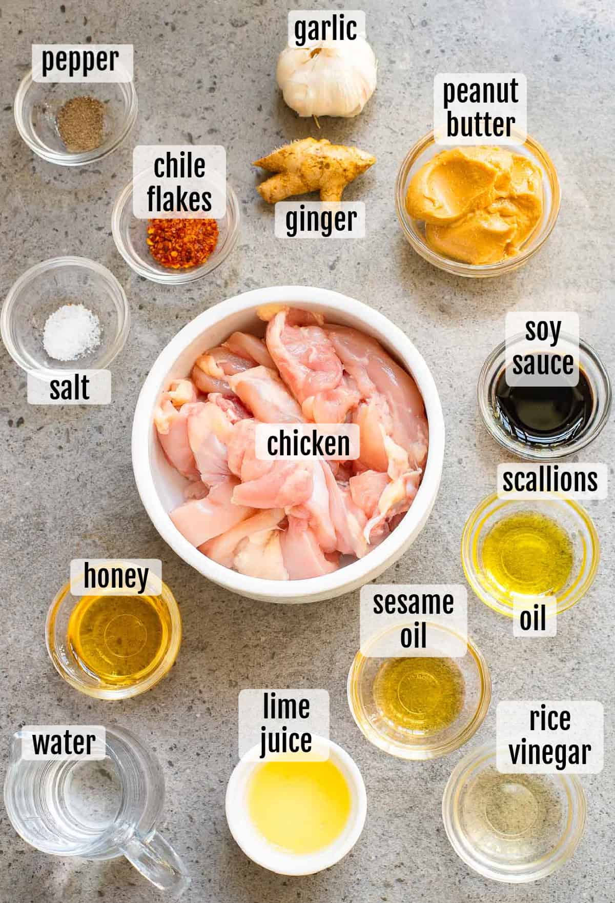 Ingredients for a recipe are arranged on a surface, including chicken, garlic, ginger, peanut butter, soy sauce, scallions, sesame oil, oil, rice vinegar, lime juice, honey, water, salt, pepper, and chile flakes.