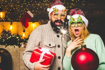 A man and woman in festive sweaters and glasses hold a sparkler, present, and large ornament. They wear Santa hats, standing in a decorated, illuminated room.