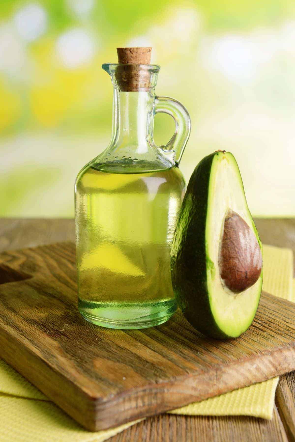 A glass bottle of avocado oil sits next to a halved avocado on a wooden board, with a blurred green and yellow background.