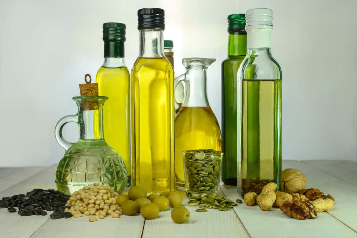 A variety of oil bottles on a wooden surface, surrounded by seeds, nuts, and olives.
