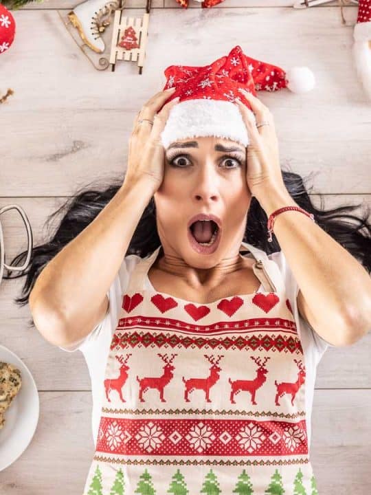 A woman wearing a Santa hat and festive apron looks surprised amidst an array of easy holiday dishes, desserts, and decorations on the table.
