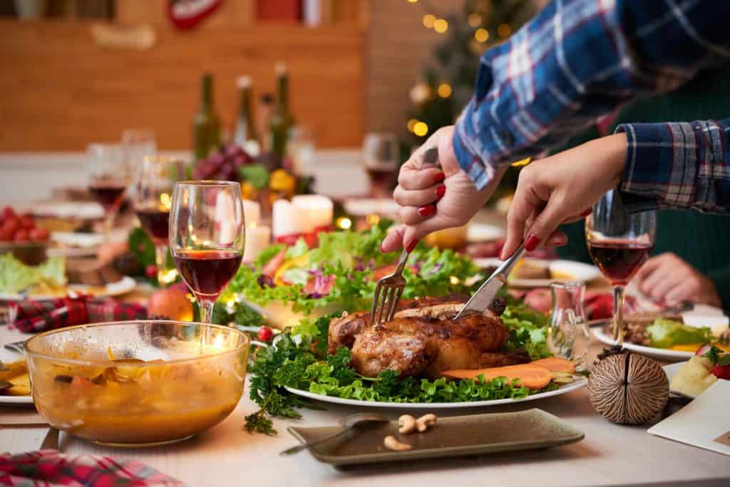 Two hands carve a roast chicken on a festive table set with wine, salad, and various dishes. Candles and Christmas decorations are in the background.