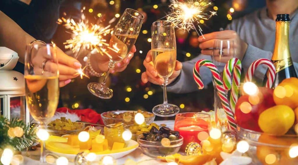 People holding sparklers and champagne glasses at a festive table with candy canes, fruits, and candles; bokeh lights in the background.