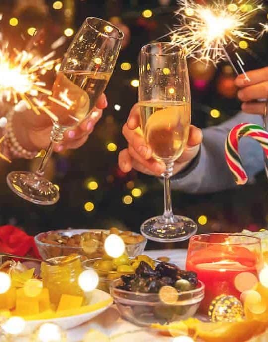 People holding sparklers and champagne glasses at a festive table with candy canes, fruits, and candles; bokeh lights in the background.