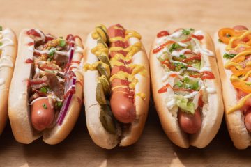 Five hot dogs with various toppings including onions, pickles, sauces, and peppers, arranged on a wooden surface.