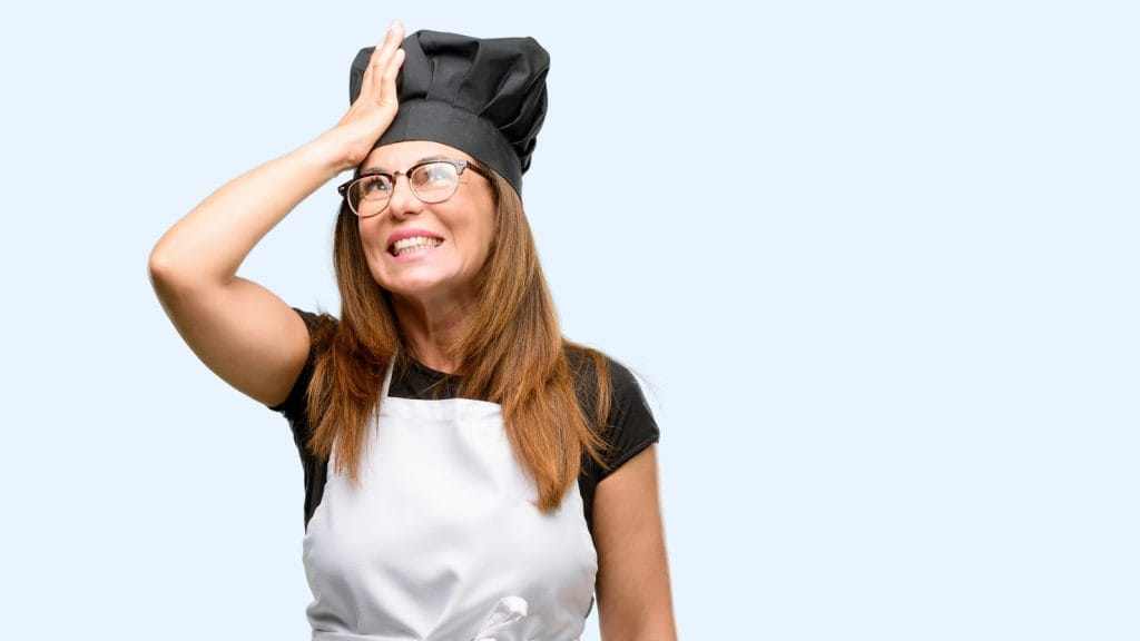 A woman wearing a chef's hat and apron holds her forehead with a smile against a light blue background.