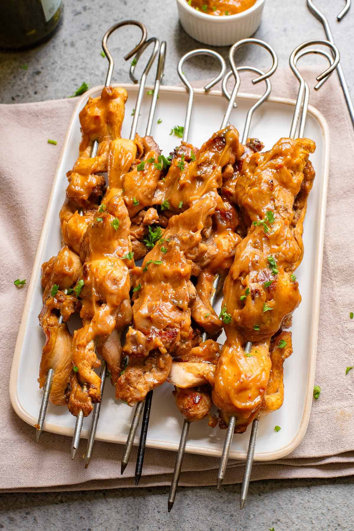 Grilled chicken skewers with a browned marinade on a white rectangular plate, garnished with parsley, with a bowl of dipping sauce in the background.