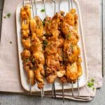 Grilled chicken skewers on a white plate, garnished with parsley, placed on a beige cloth.