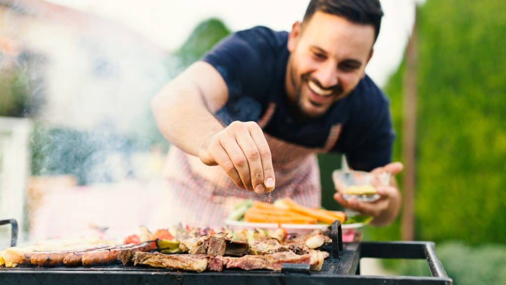 A man sprinkles seasoning over meat on a barbecue grill, smiling with vegetables on the side.