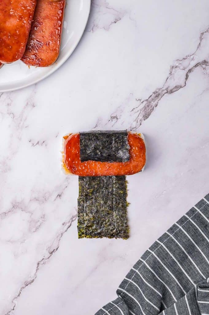 A piece of Spam musubi with a slice of meat on rice, wrapped in nori, is placed on a marble surface. A plate with additional slices is partially visible.