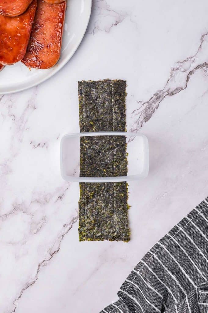 Nori sheet positioned on a marble surface with a plastic container on top; slices of meat are partially visible on a white plate in the corner.