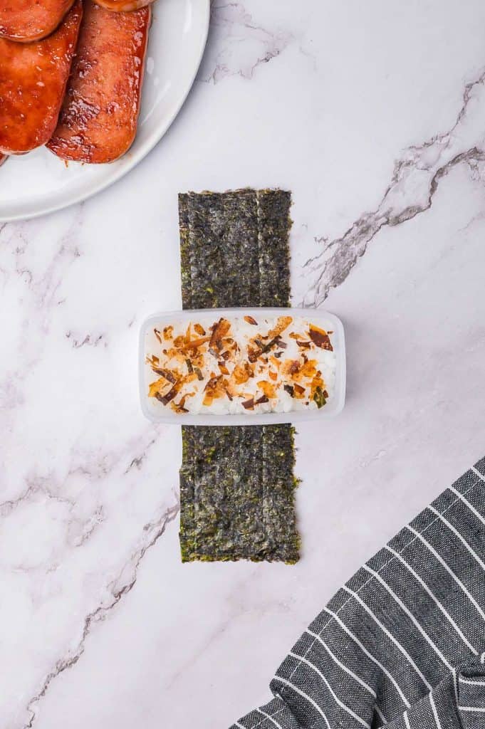 A plate with cooked meat next to a dish of rice topped with seaweed flakes placed on a strip of seaweed on a marble surface, with a striped cloth in the corner.