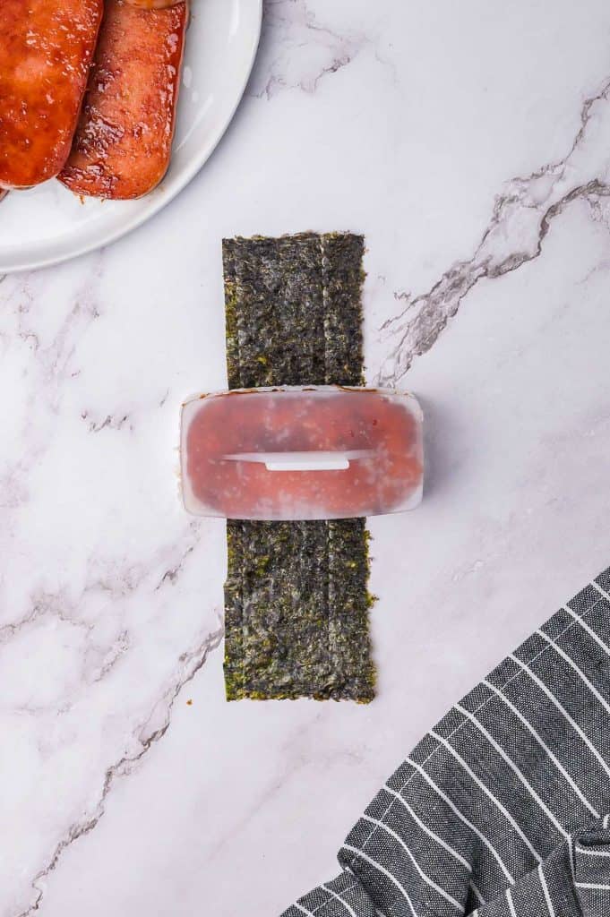 A block of luncheon meat placed on top of rice and seaweed on a counter, with a plate of additional slices nearby.
