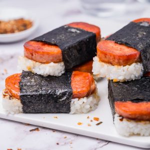 Close-up of a plate with multiple pieces of spam musubi, featuring slices of spam on rice, wrapped with seaweed, on a white surface.