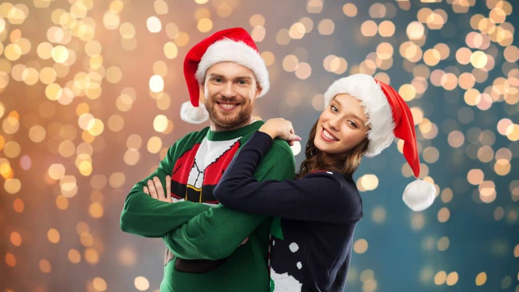 A man and woman wearing Santa hats smile in festive sweaters against a bokeh background.