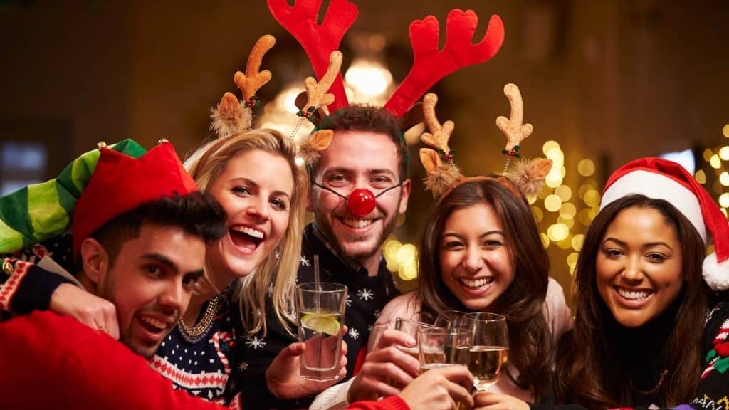 Five people wearing festive holiday attire and antlers are smiling and holding drinks in a warmly lit setting.