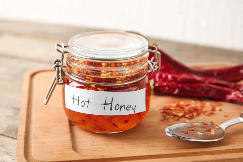 A glass jar labeled "Hot Honey" on a wooden board, with dried chili peppers and a spoon with chili flakes beside it.