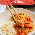 A bowl of white rice topped with stir fried tomatoes and eggs, garnished with green onions, being picked up with chopsticks.