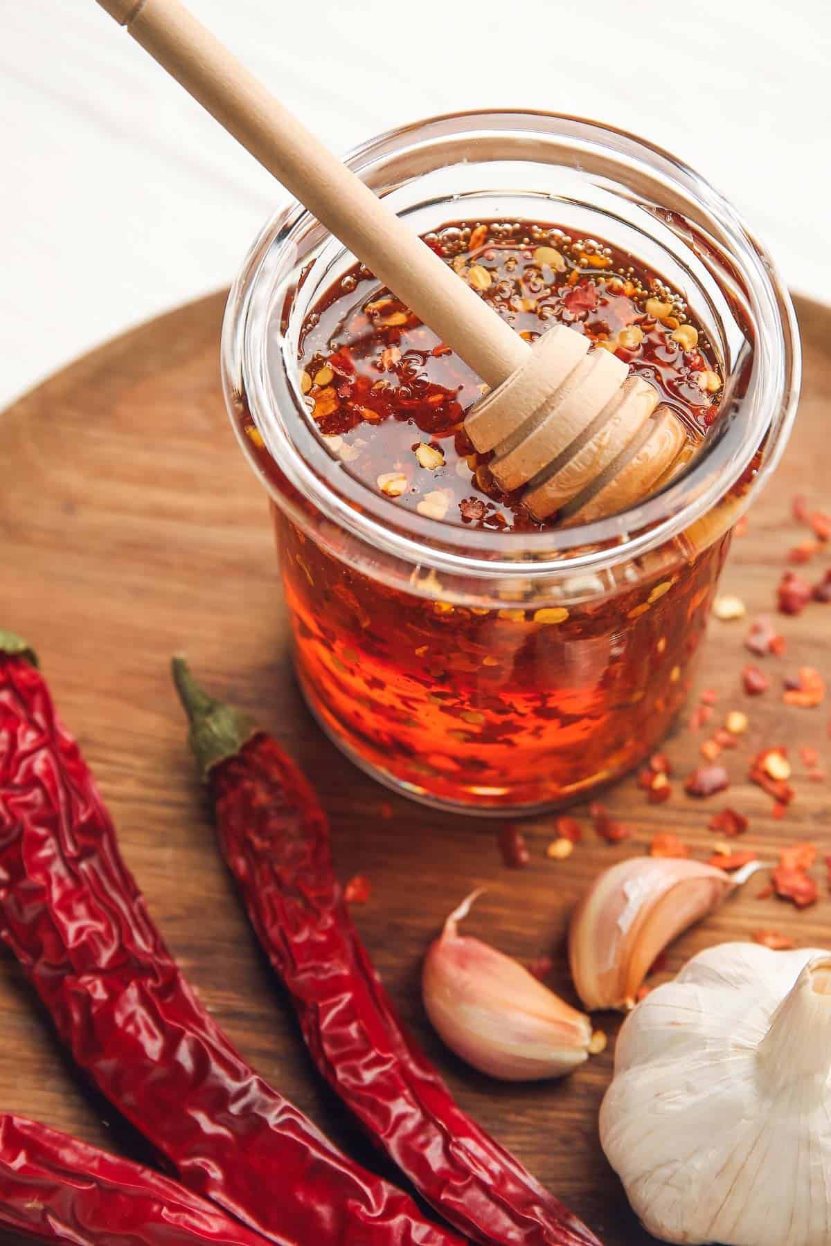 Glass jar of chili-infused honey with a dipper on a wooden board, surrounded by dried red chilies and garlic cloves.