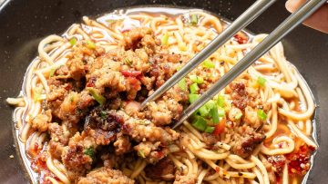 A bowl of noodles topped with minced meat, chopped green onions, and chili peppers. Chopsticks are picking up some noodles.