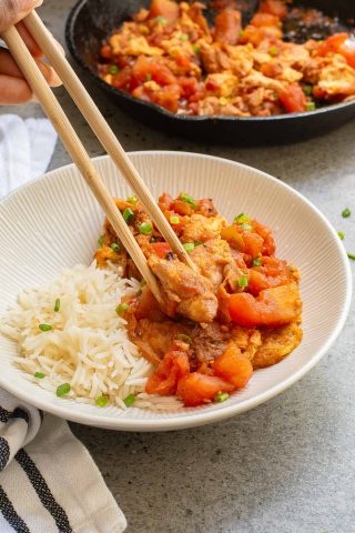 A bowl containing white rice and stir fried tomatoes and eggs, with chopsticks lifting a piece of chicken. A skillet with more of the dish is in the background.