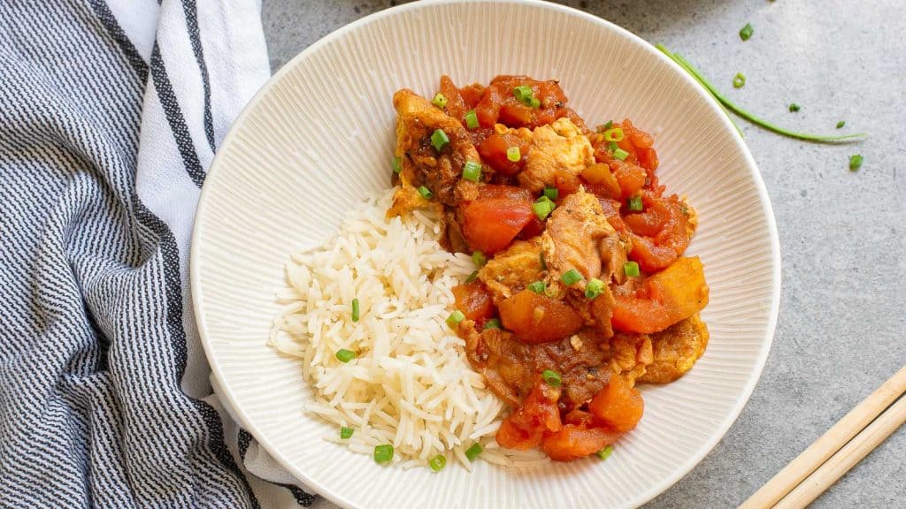 A white plate with rice and stir fried tomaotes and eggs, garnished with green onions. A striped towel is partially visible on the side.