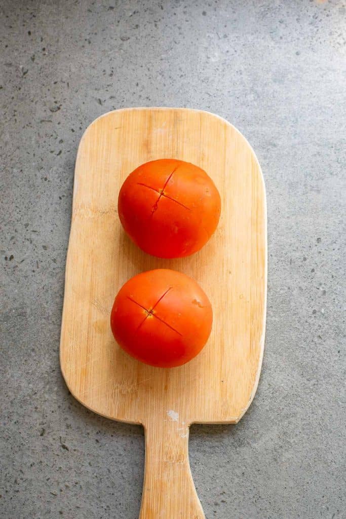 Two tomatoes with cross-shaped cuts on top, placed on a wooden cutting board on a gray surface.