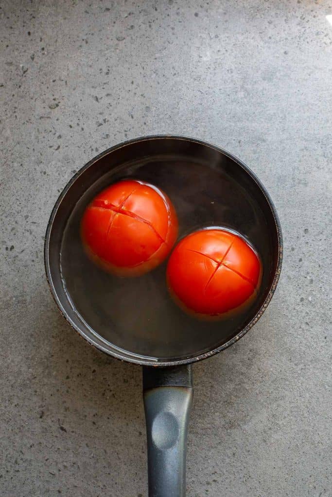 Two tomatoes with cross-shaped cuts on top sit in a small saucepan filled with water.