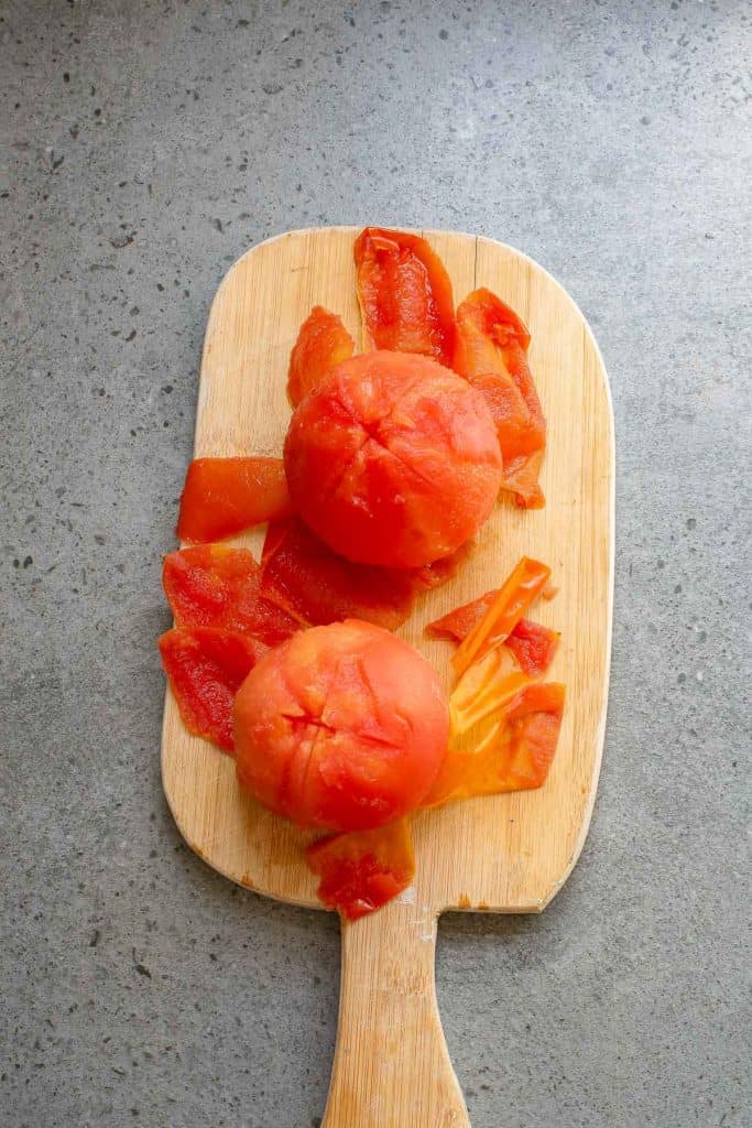Peeled tomatoes on a wooden cutting board with tomato skins placed around them.
