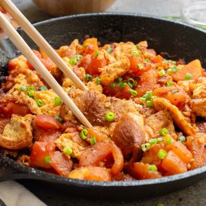 A skillet filled with stir fried tomatoes and eggs with green onions. Chopsticks are used to serve the dish.