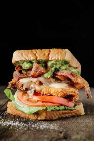 A sandwich with toasted bread, lettuce, tomato, fried chicken, melted cheese, bacon, and guacamole on a dark background.