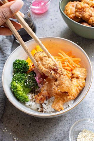 A hand uses chopsticks to hold fried chicken over a bowl filled with rice, broccoli, sliced mango, and carrots, garnished with pickled onions and sesame seeds.